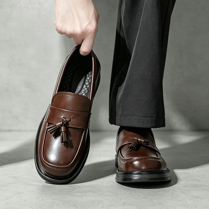 Man holding brown tassel loafer beside foot wearing matching pair